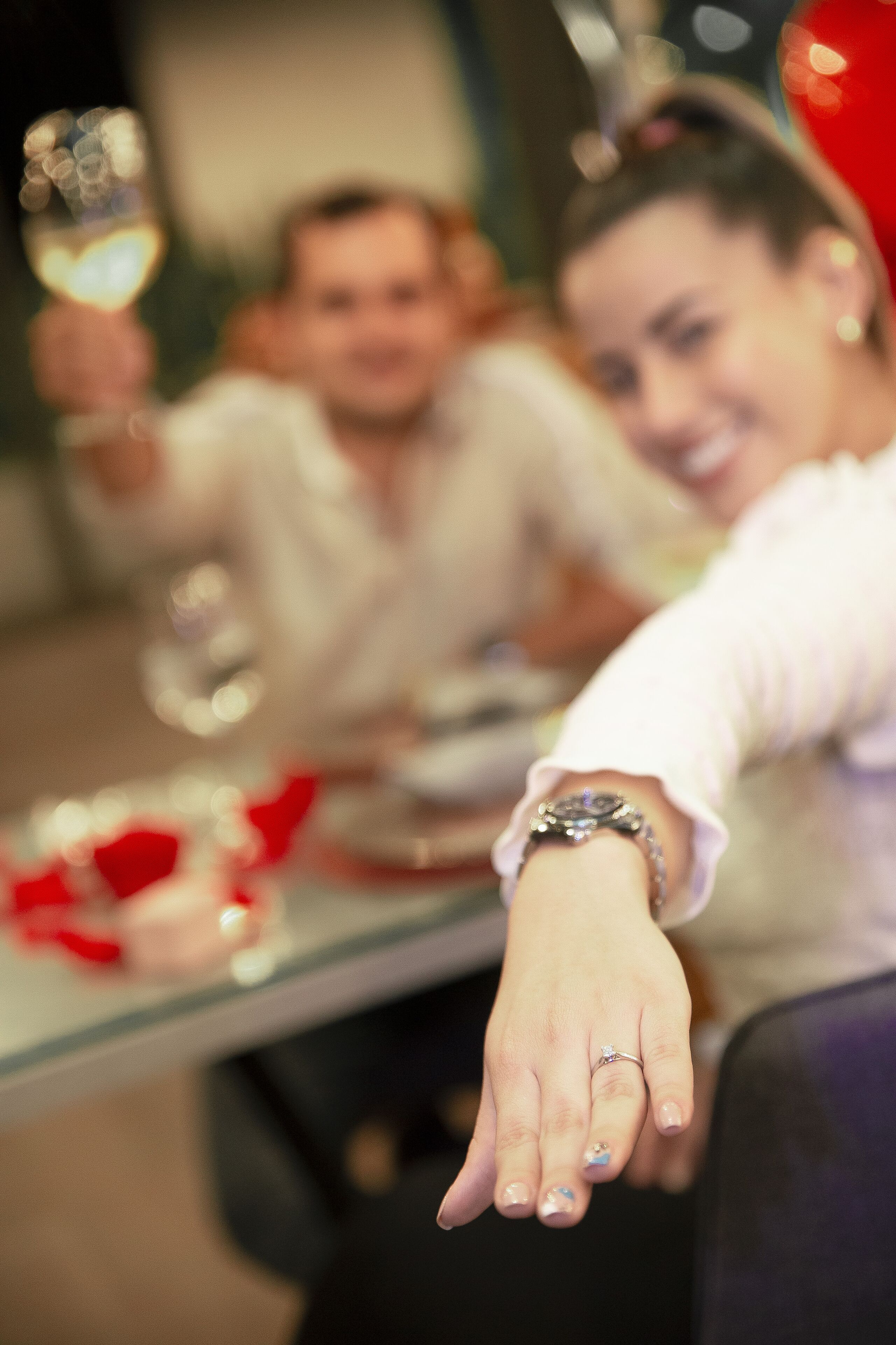 Une personne tend la main, portant une bague, et une autre personne se tient à l’arrière-plan, tenant un verre.
