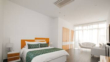 A modern hotel room with a large bed, a bedside table with a lamp, a wardrobe, a sofa, and a large window providing natural light.