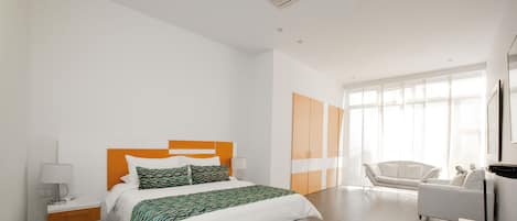 Una habitación de hotel moderna con una cama grande, una mesita de noche con lámpara, un armario, un sofá y un ventanal que deja entrar luz natural.