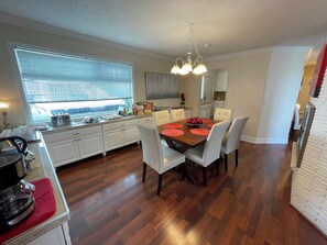 A modern kitchen with white cabinets, a wooden dining table with red placemats, and a large window with blinds. - Arctic Fox Inn (Anchorage)