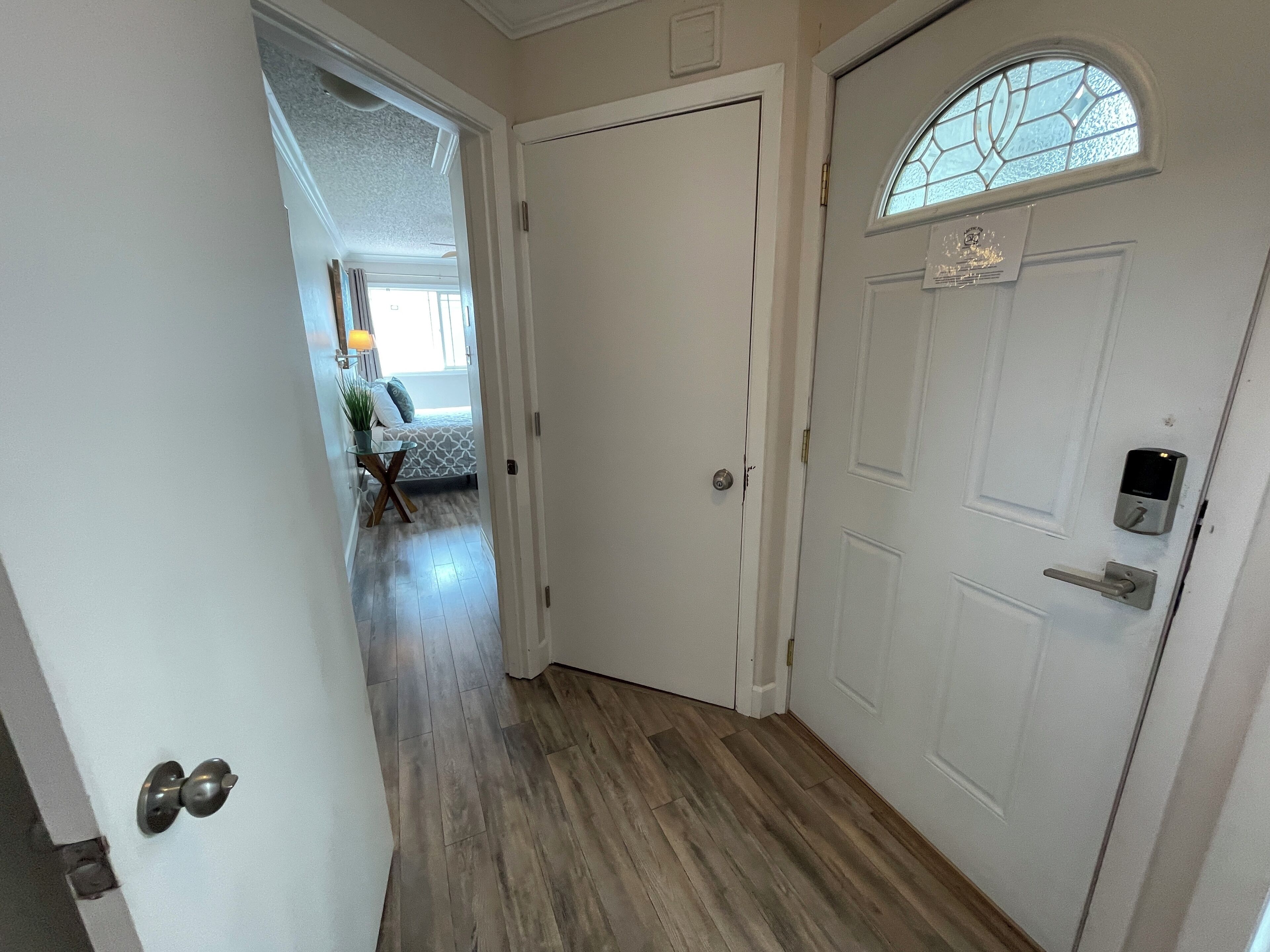 A hallway with a wooden floor, a white door with a peephole, and a small window with a decorative glass design.
