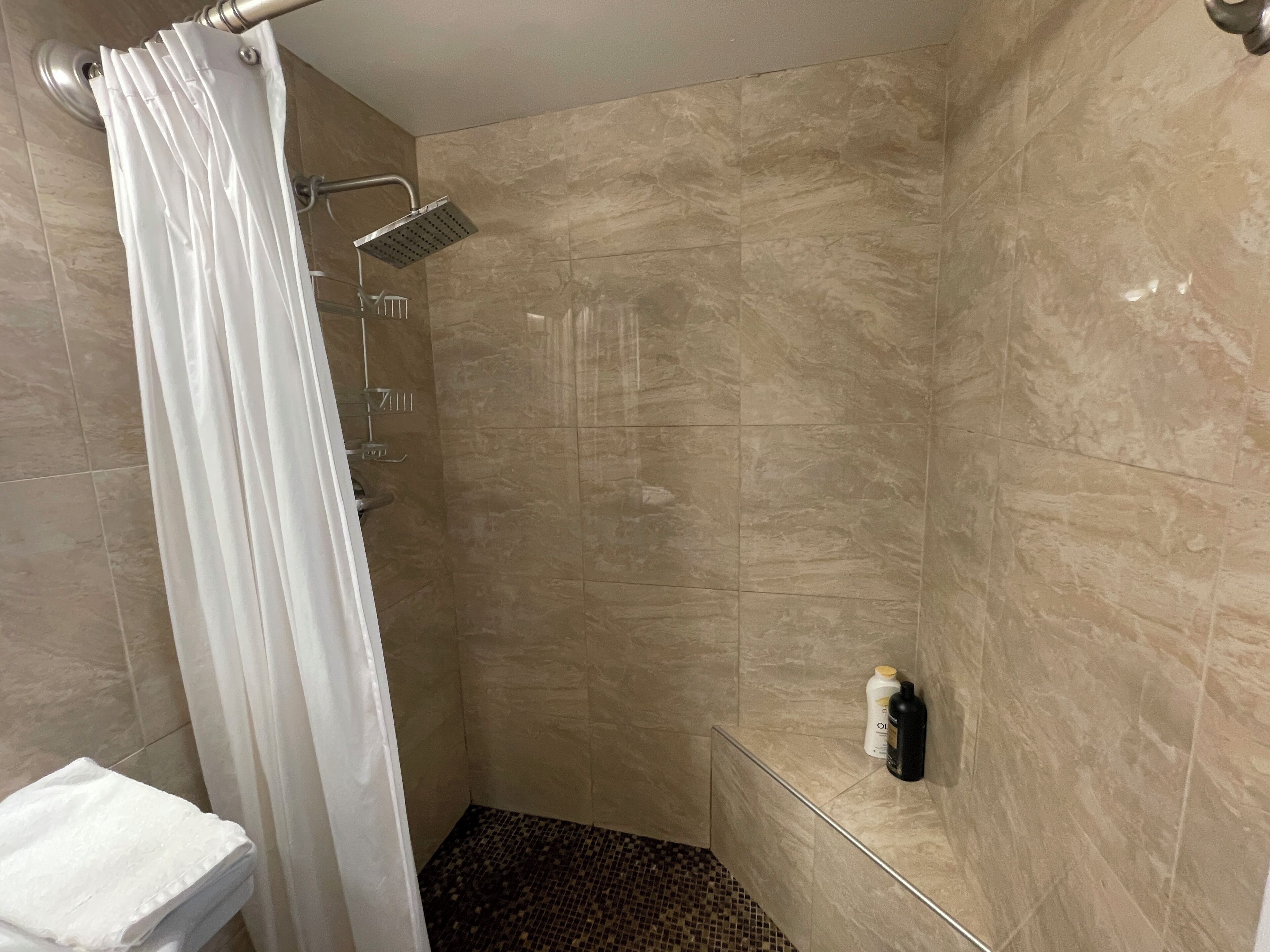 Een douche met een wit gordijn, een handdouche en een fles shampoo aan de muur.