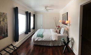 A bedroom with a bed, a ceiling fan, a window with curtains, a painting on the wall, and a small table with a lamp. - Arctic Fox Inn (Anchorage)
