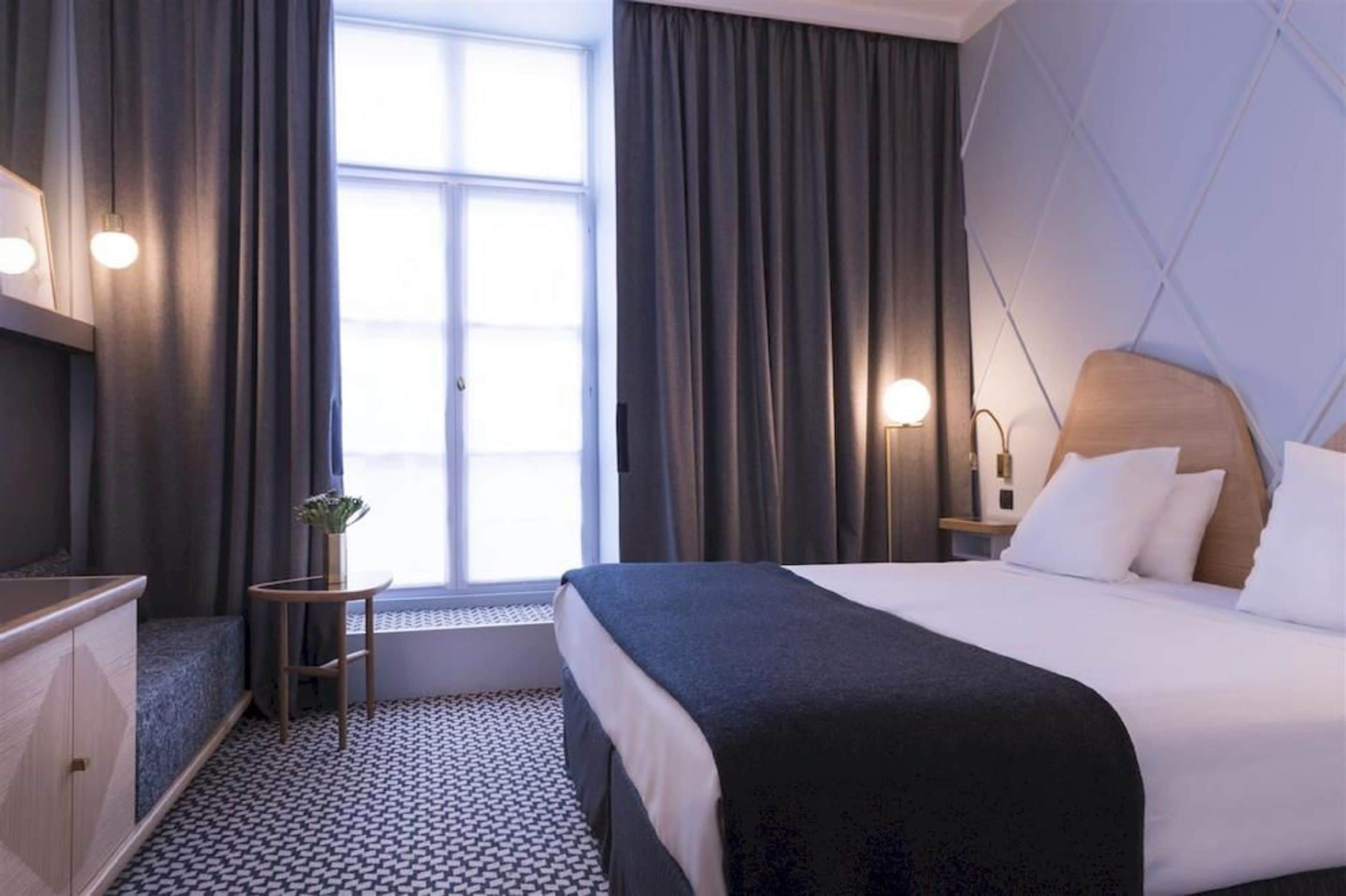 A hotel room with a bed, a desk, a chair, a small table with a plant, and a window with curtains.