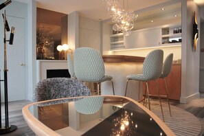 Ein modernes Wohnzimmer mit einem Glastisch, zwei gemusterten Sesseln, einem Kamin und einer Küchenzeile mit Einbauschränken.