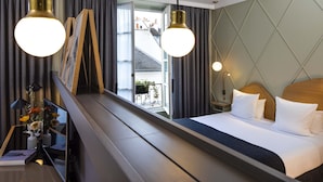 A hotel room with a bed, bedside tables, a mirror, curtains, and a view of a balcony with plants. - Millésime Hôtel (Paris)