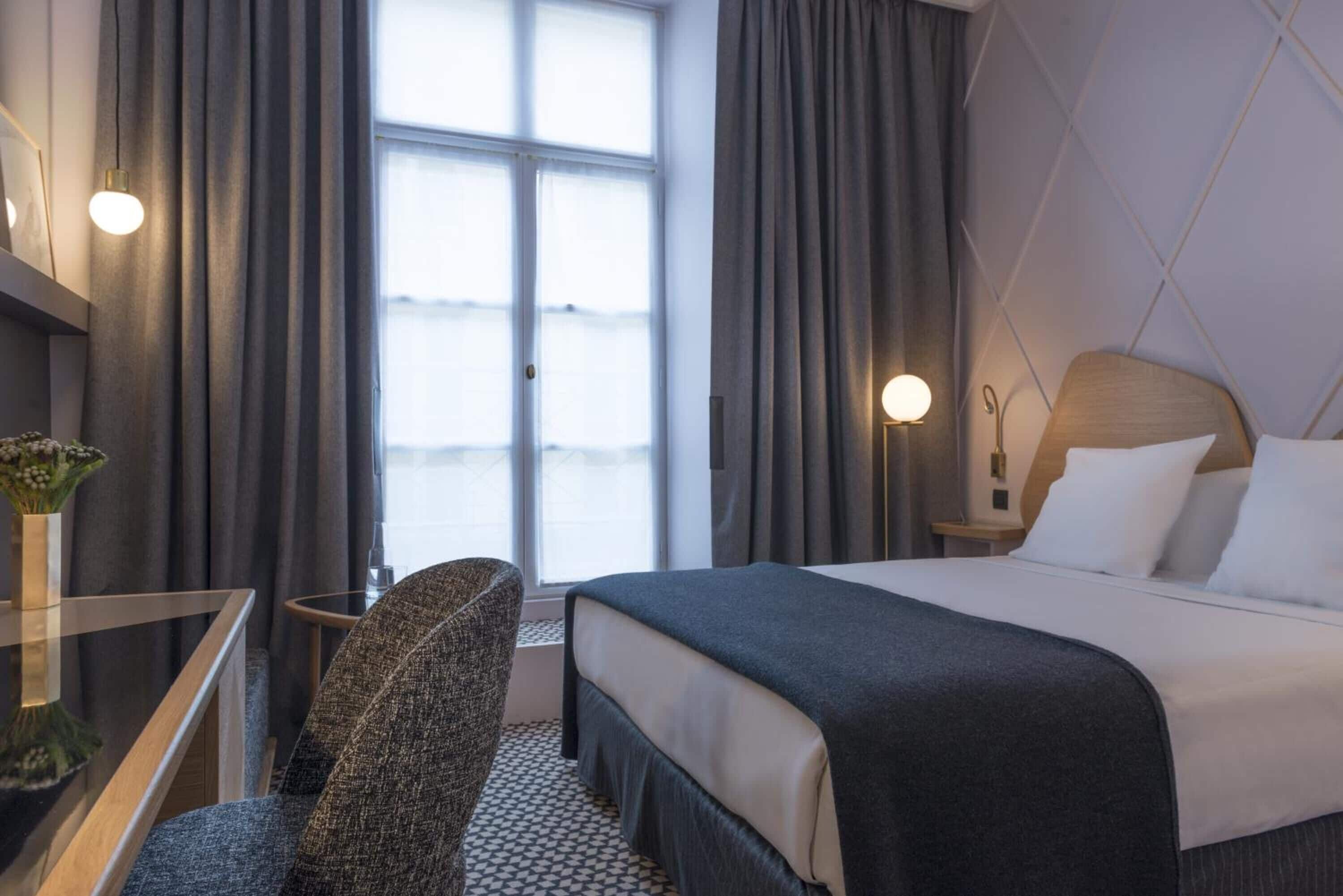 Quarto de hotel com cama, escrivaninha, cadeira, janela com cortinas e luminária na parede.