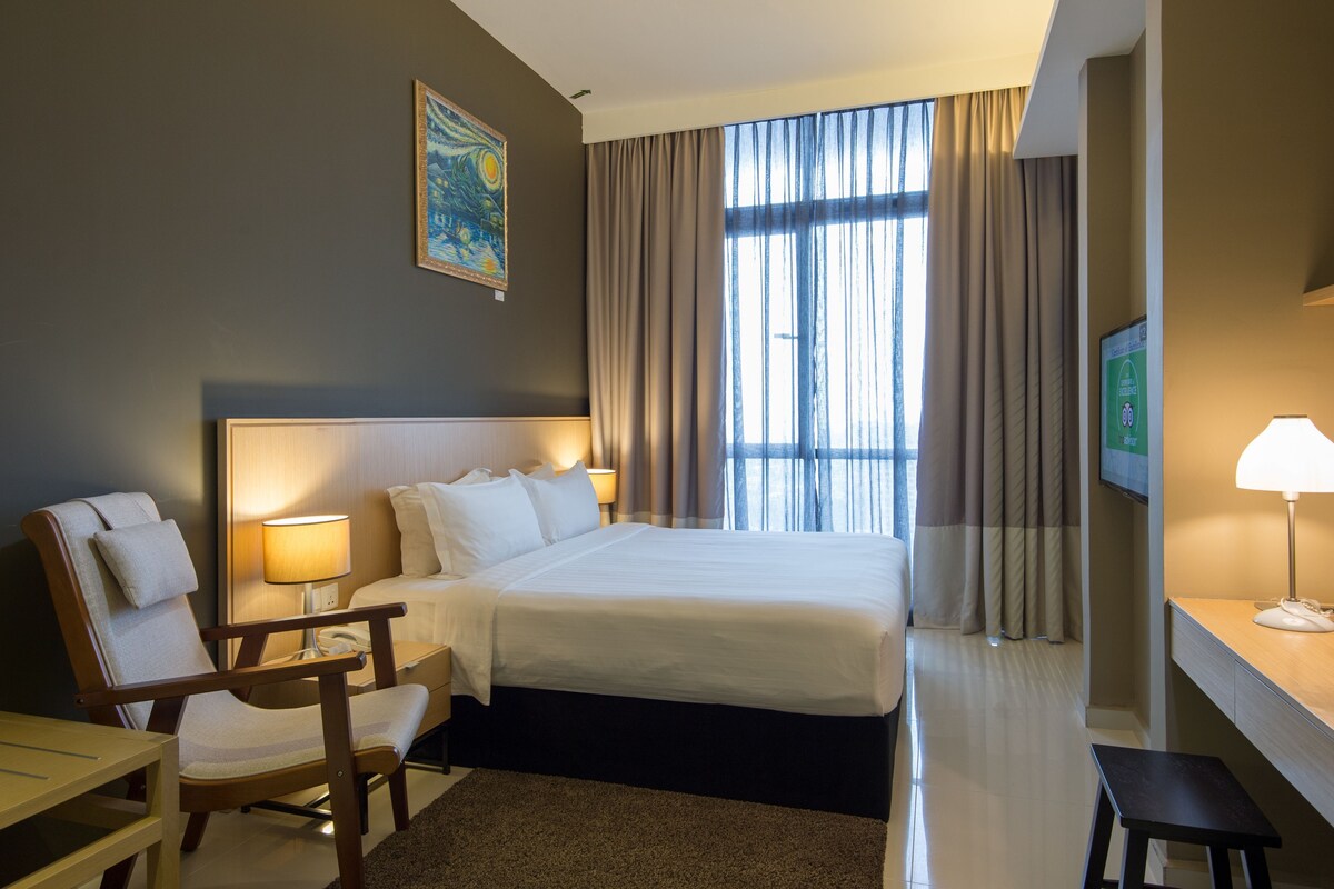 Habitación de hotel con cama, escritorio, silla, lámpara, un cuadro en la pared y un ventanal con cortinas.