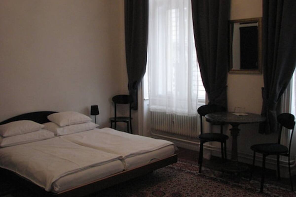 Un dormitorio con cama, una mesita, dos sillas y una ventana con cortinas.