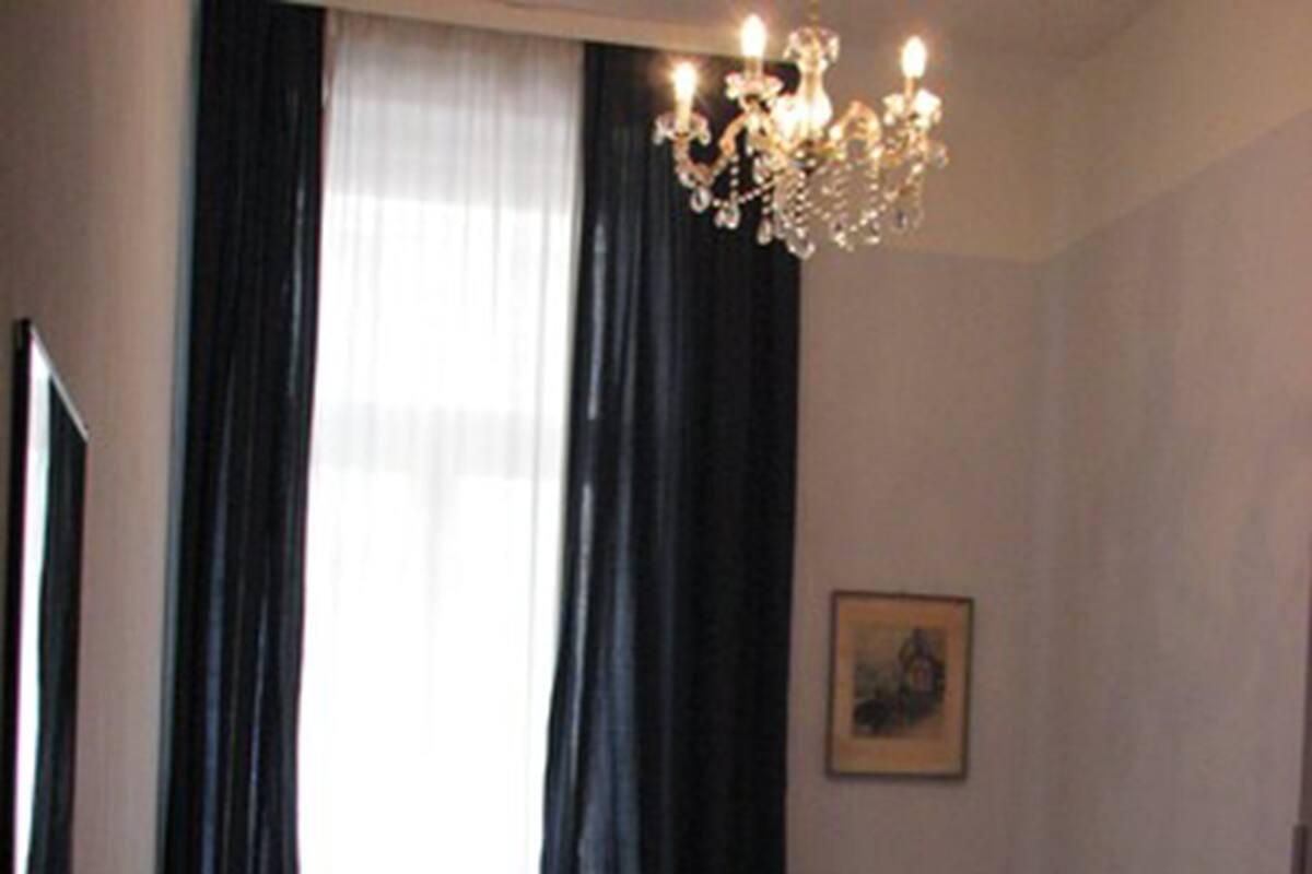 Habitación de hotel con cama, escritorio, silla, pequeña mesa, lámpara, candelabro y un cuadro en la pared.