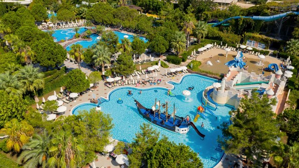 Vue aérienne d’un complexe hôtelier doté de nombreuses piscines, dont une grande piscine centrale sur le thème d’un navire de pirates, entourée d’une végétation luxuriante et de diverses attractions aquatiques.