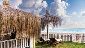 Un espace aménagé au bord de la plage, avec des parasols en chaume, des chaises longues et une clôture en bois donnant sur l’océan.
