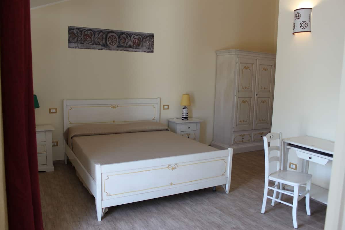 A bedroom with a bed, a desk, a chair, a wardrobe, and a nightstand with a lamp.