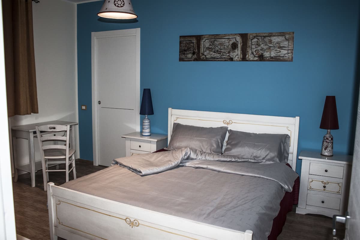 A bedroom with a bed, a desk, a chair, a nightstand, and a wall art piece.
