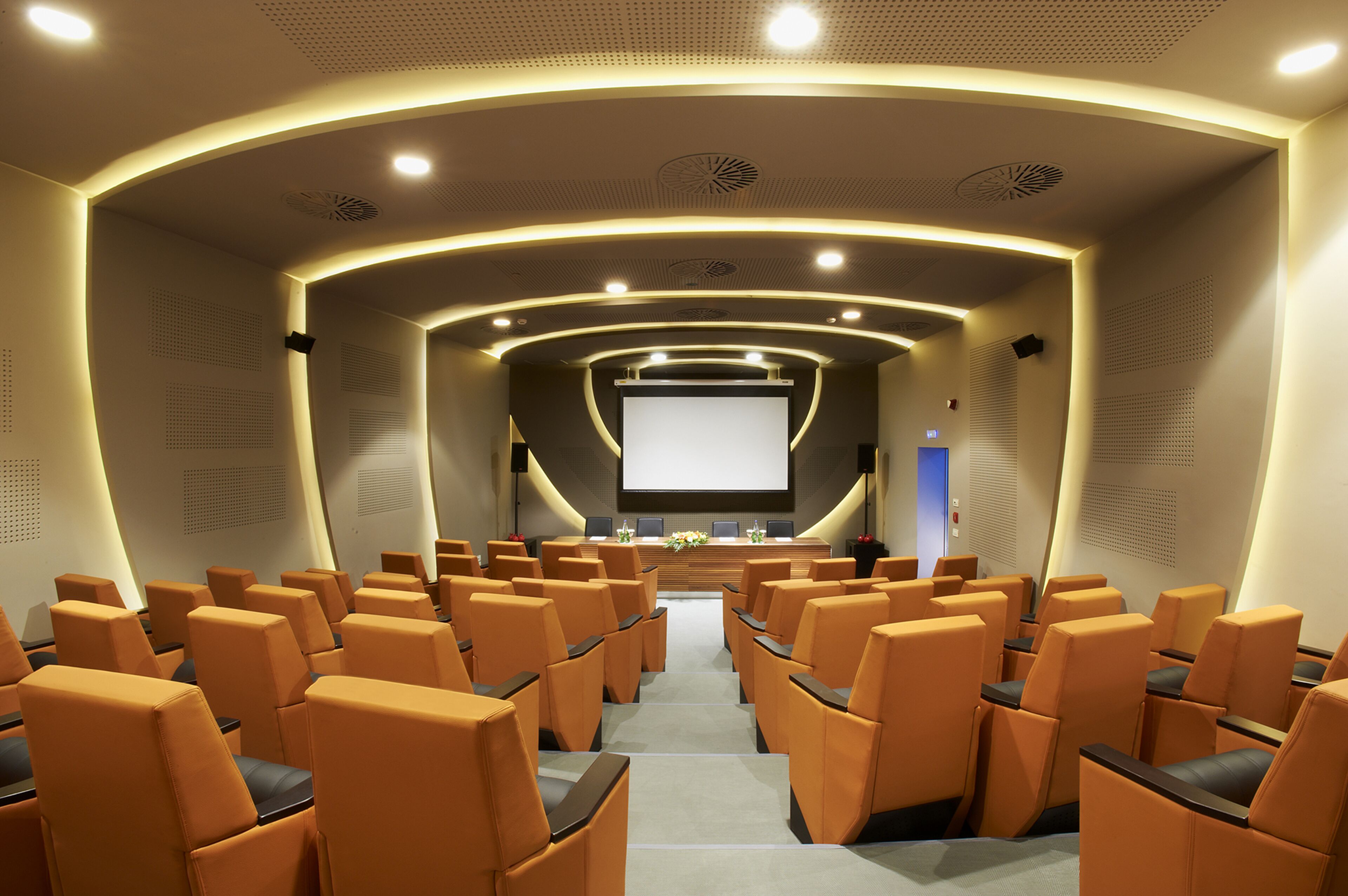 A conference room with rows of orange chairs, a large screen at the front, and a podium with a floral arrangement.