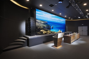 Uma sala de conferências moderna com uma tela grande exibindo uma paisagem cênica, um balcão de recepção e uma escadaria que leva aos níveis superiores.