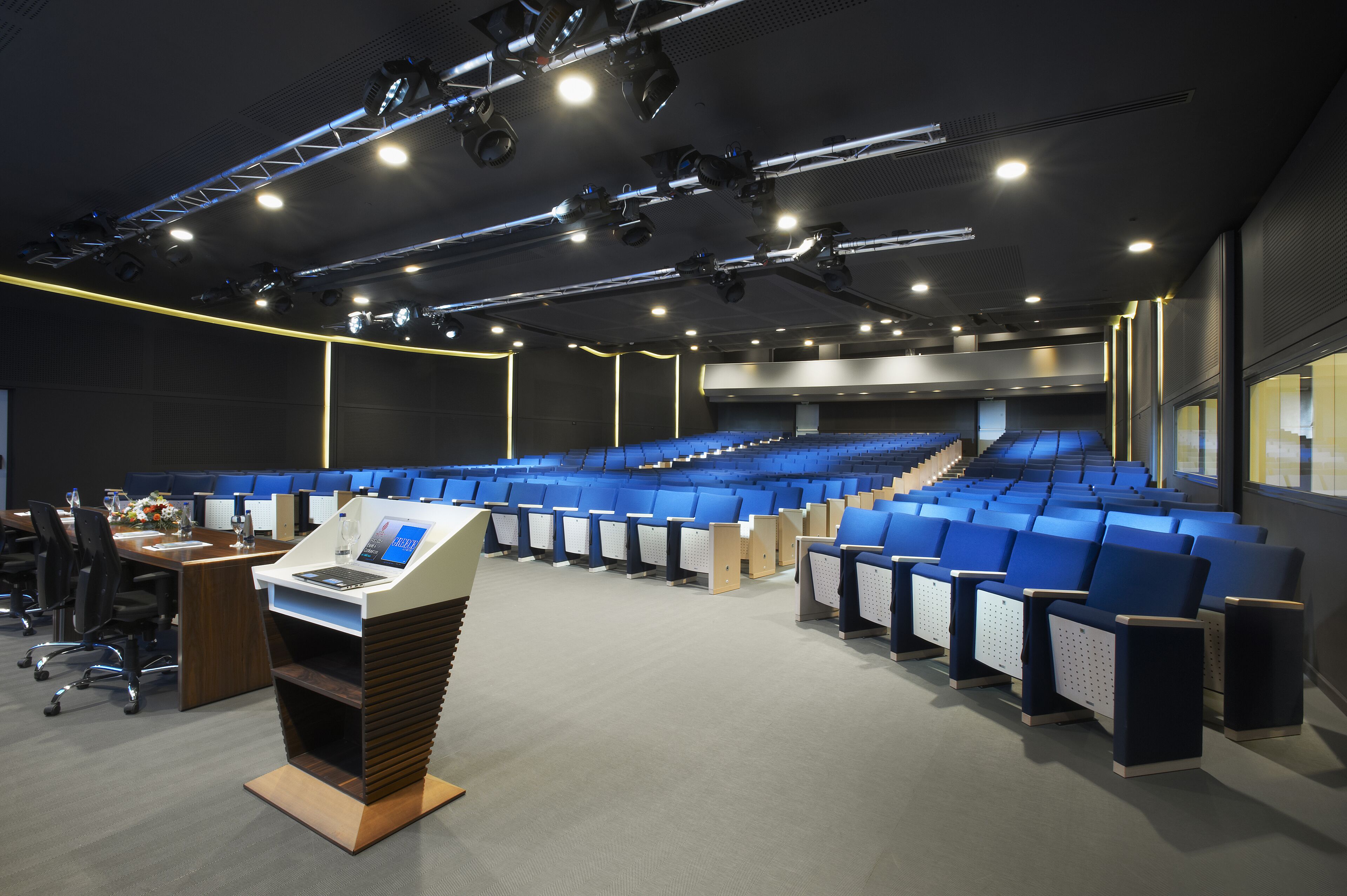 Konferenssisali, jossa on riveissä sinisiä tuoleja, puhujapönttö ja valaistuslaitteilla varustettu lava.