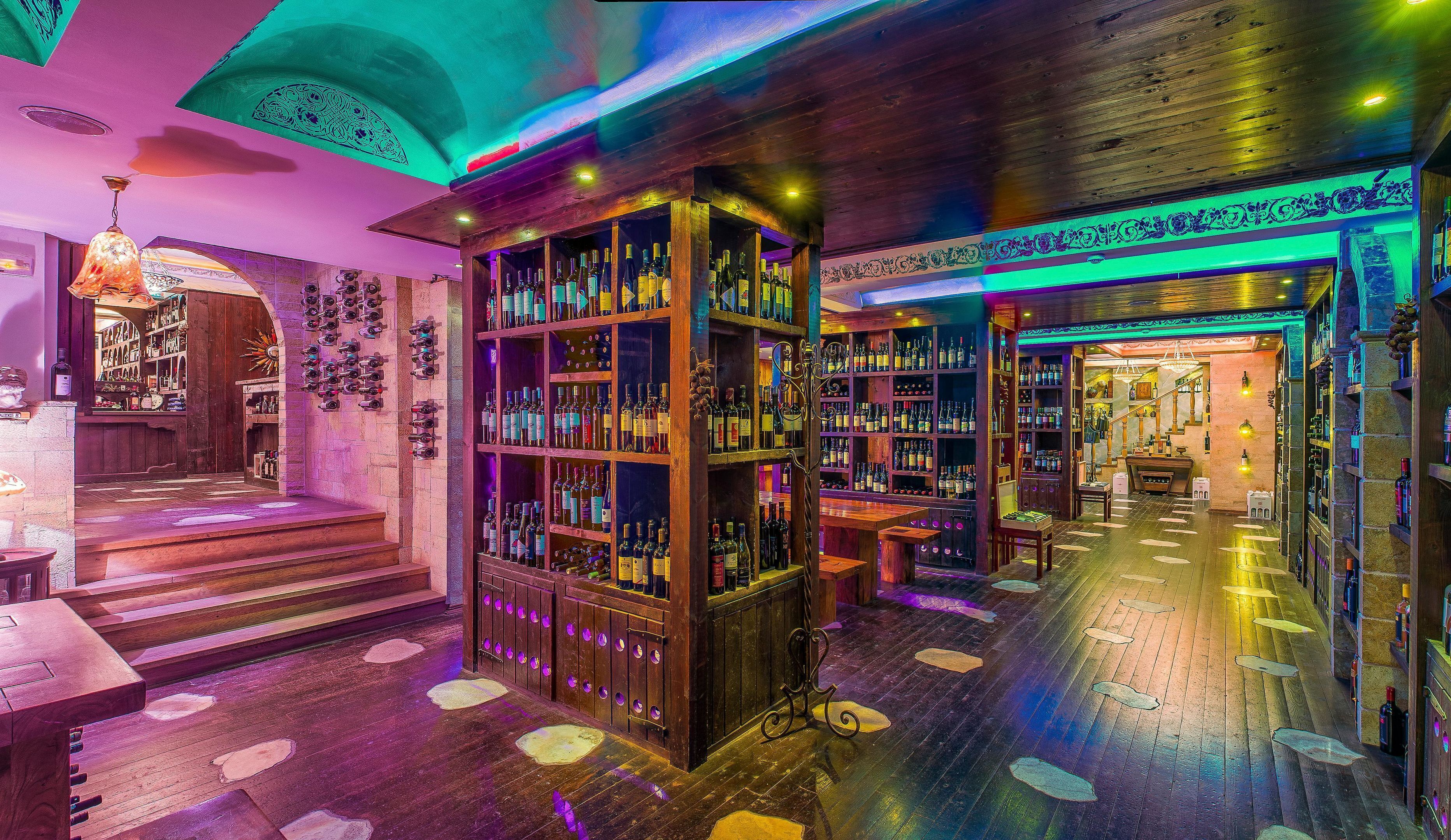 A bar area with wooden shelves stocked with bottles, a staircase leading to a seating area, and a decorative ceiling.