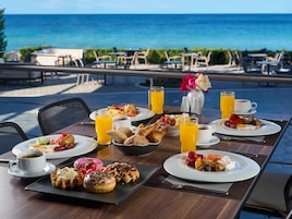 Mesa de café da manhã com diversos pratos, bebidas e vista para o oceano.