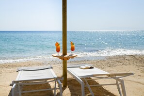 Escena de playa con dos sillones de descanso, una mesa con un libro y dos bebidas sobre un soporte de madera.