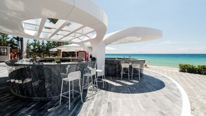 A modern outdoor bar area with a curved marble countertop and white bar stools, overlooking a beach and the sea. - Potidea Palace Hotel - All inclusive (Nea Propontida)