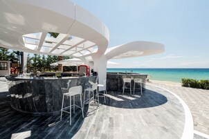 Uma área de bar moderna ao ar livre, com bancada de mármore curva e banquetas brancas, com vista para uma praia e o mar.