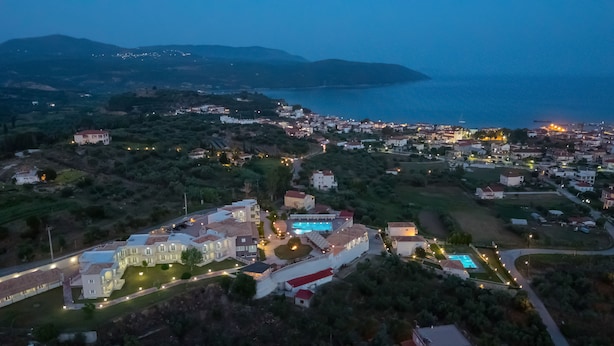 Vista aérea de un pueblo costero al anochecer, con edificios iluminados, una piscina y un paisaje montañoso al fondo.