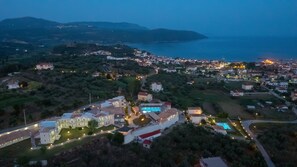 An aerial view of a coastal town at dusk with illuminated buildings, a swimming pool, and a hilly landscape in the background. - Panorama Resort (Pylos-Nestoras)