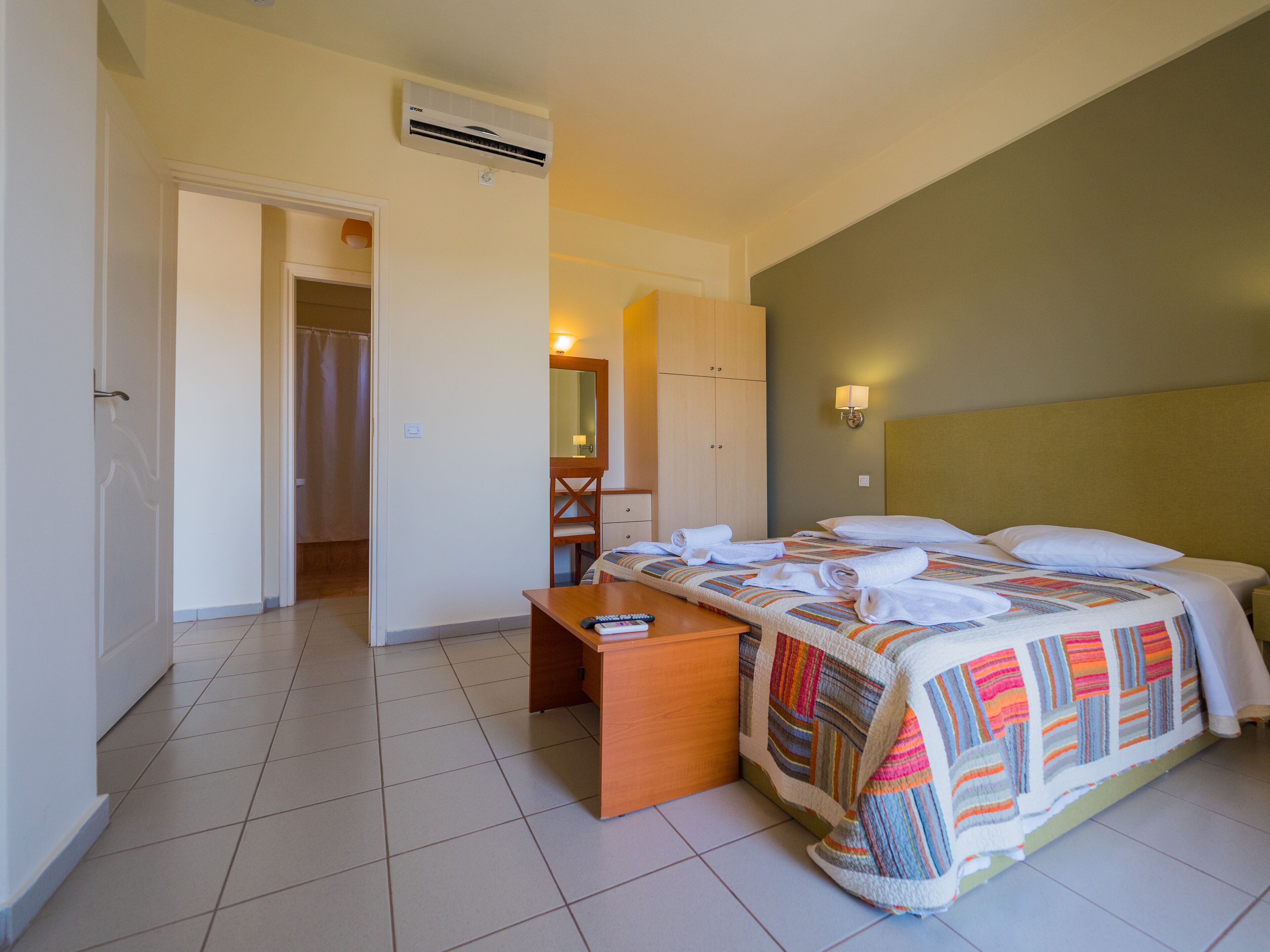 Habitación de hotel con cama, escritorio, espejo y baño visible a través de una puerta abierta.