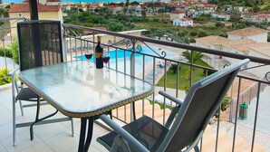 Un balcón con mesa y sillas de vidrio, con vista a una piscina y un pueblo costero.