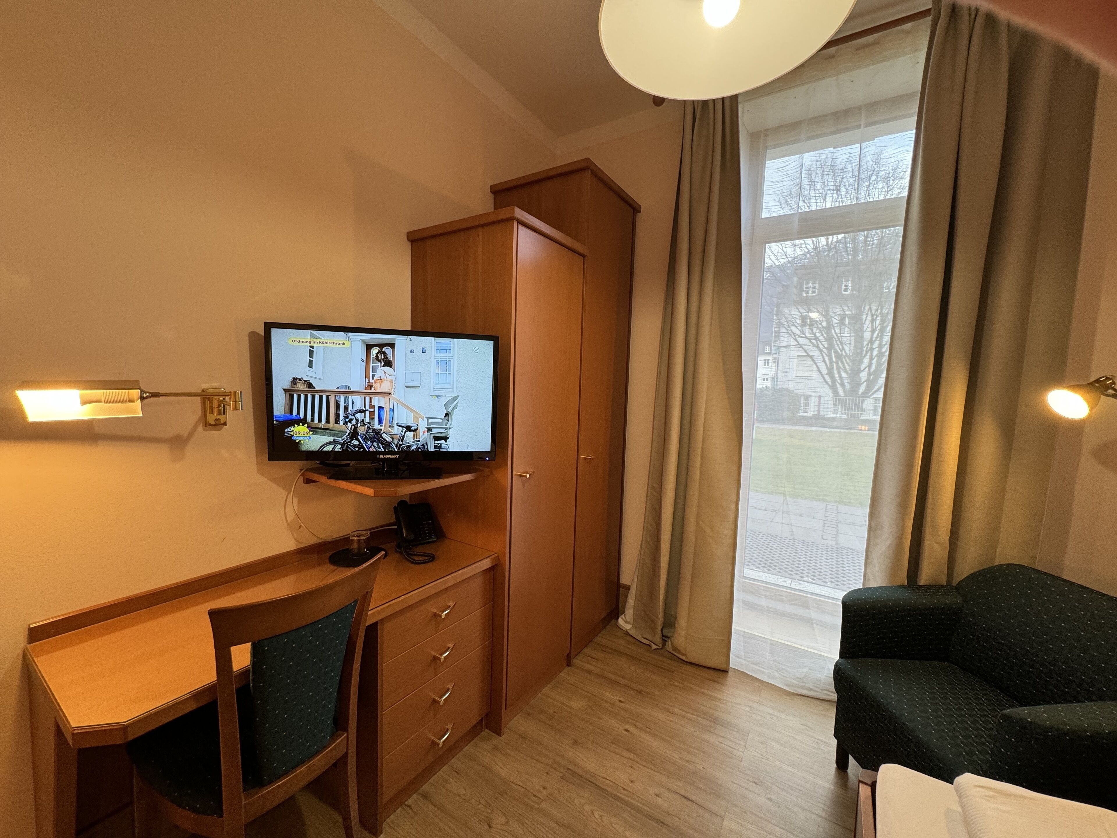 Une chambre d’hôtel avec un bureau en bois, un téléviseur fixé au mur, une armoire, un canapé et une grande fenêtre avec des rideaux.