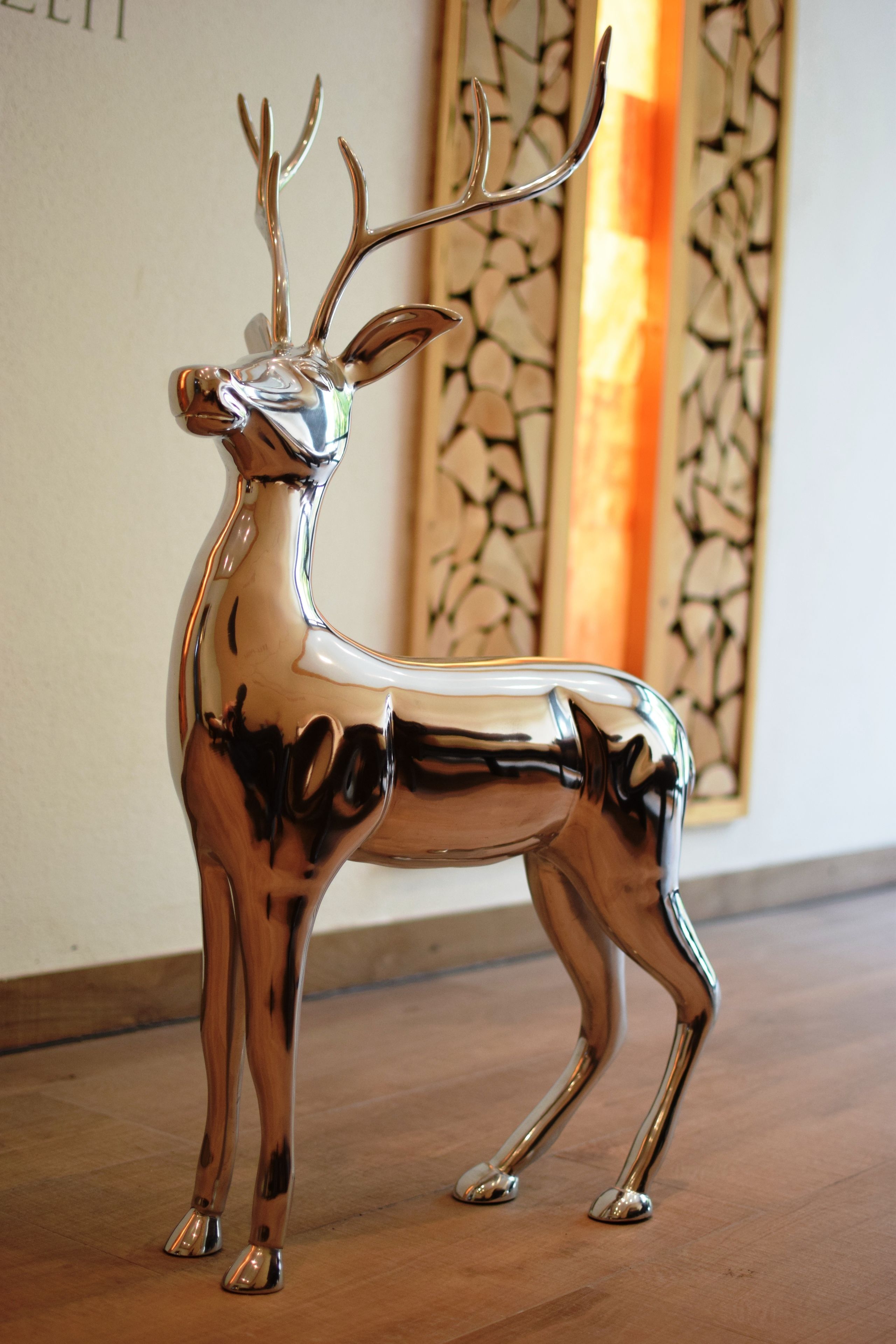 A metallic deer sculpture on a wooden floor with a decorative wall panel in the background.