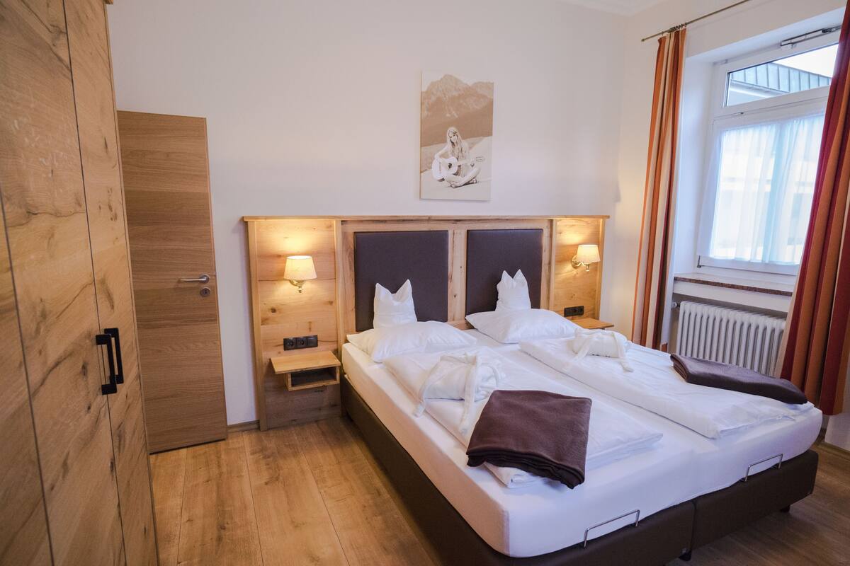 Una habitación de hotel con un armario de madera, una cama con ropa de cama blanca, dos mesitas de noche con lámparas y una ventana con cortinas.
