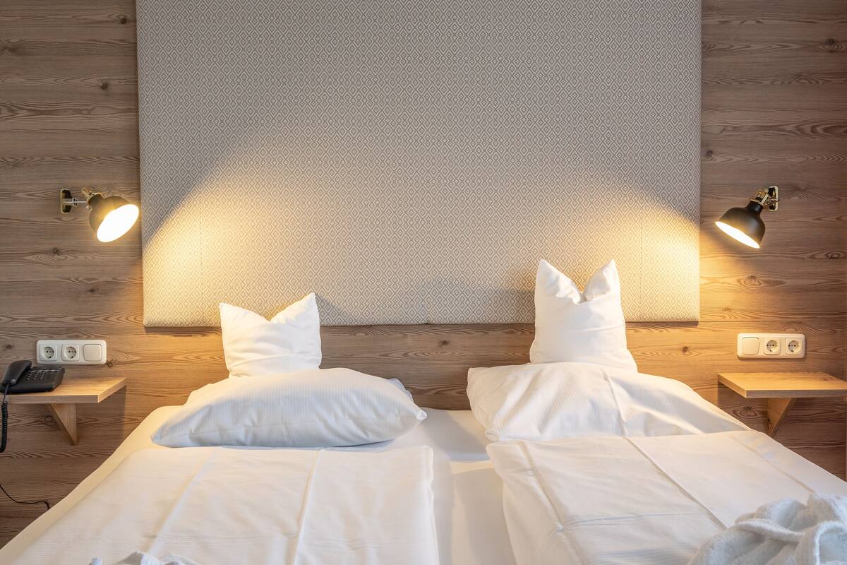 Una cama doble con sábanas y almohadas blancas, dos mesitas de noche con lámparas, y un aplique en la pared.