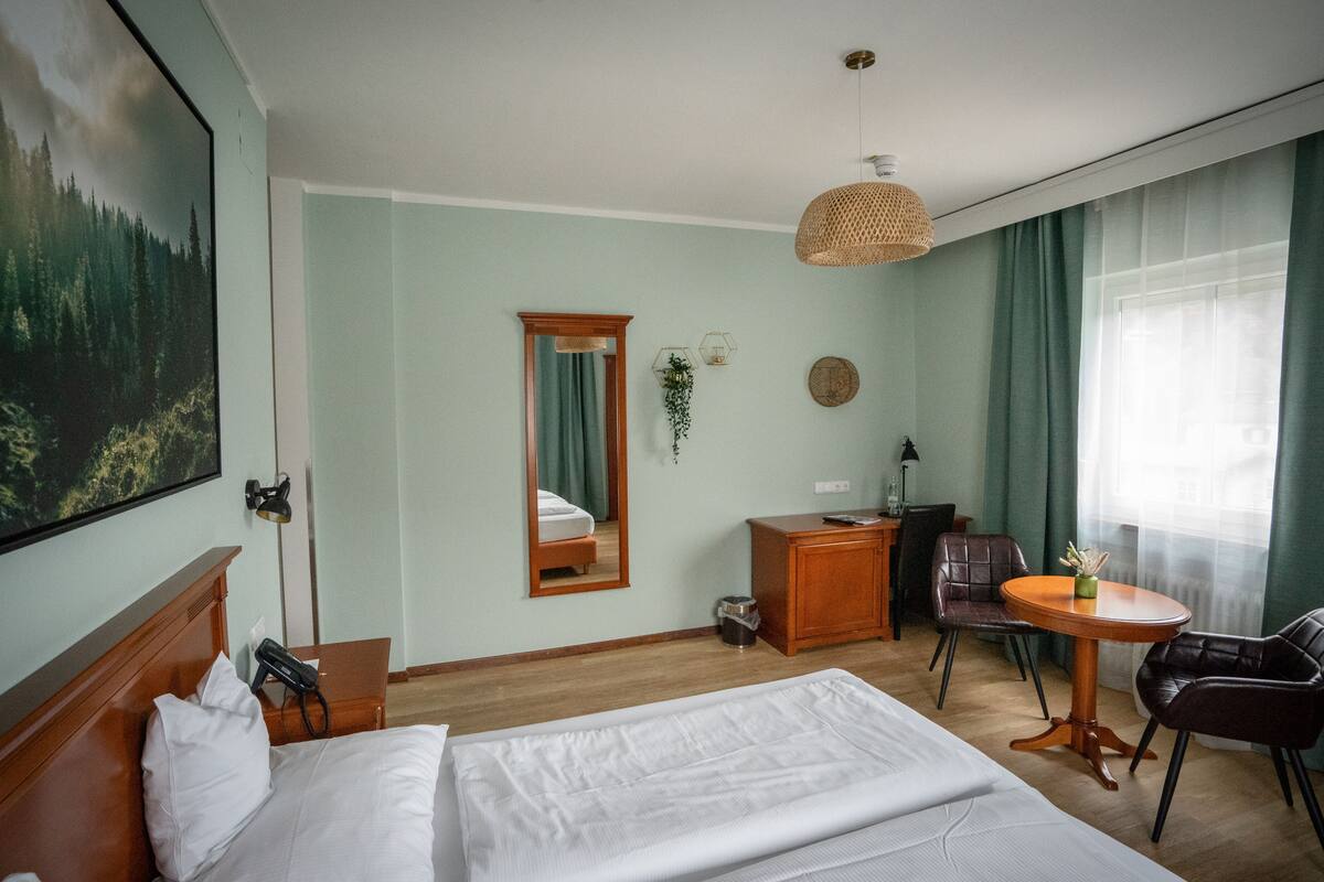 Habitación de hotel con cama, escritorio, silla, mesita, espejo y una ventana grande con cortinas.