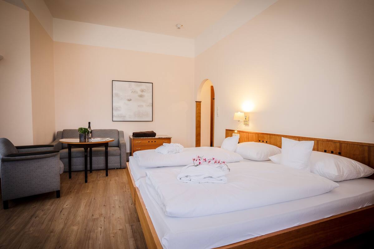 Habitación de hotel con cama de madera, sofá gris, una mesita con una planta y una lámpara fijada a la pared.