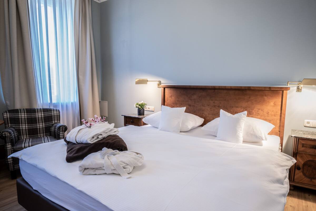 Una cama bien hecha con sábanas y almohadas blancas, un sillón escocés, una mesita de noche de madera con una lámpara y una ventana con cortinas traslúcidas.