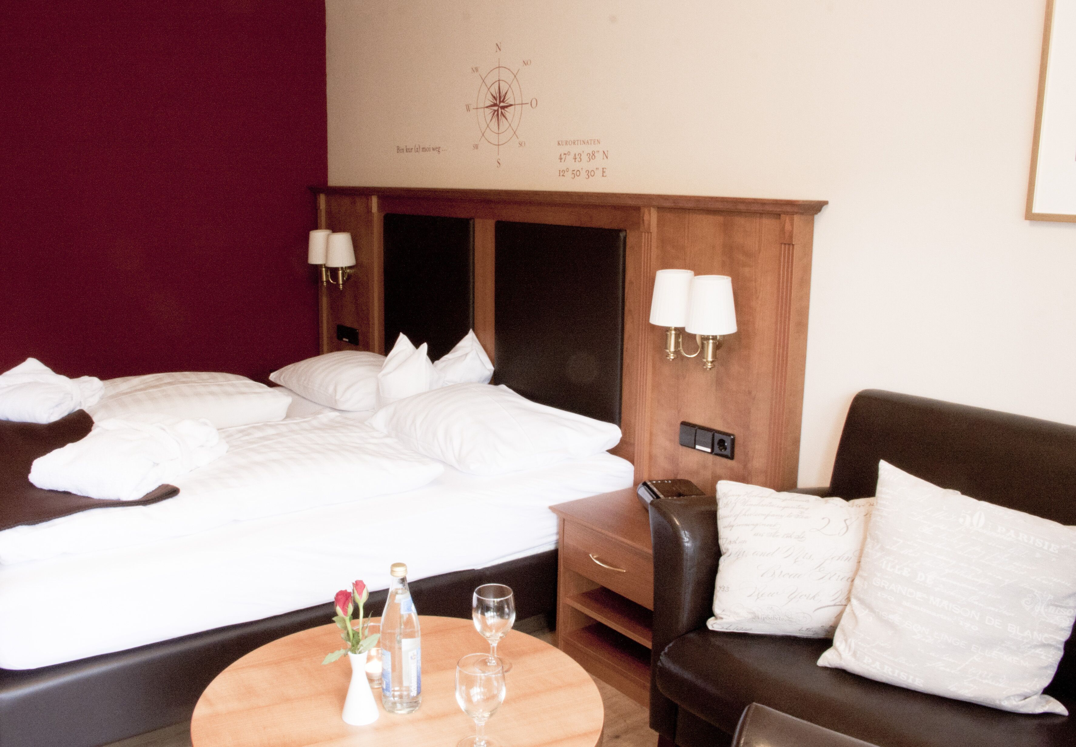A hotel room with a large bed, a wooden headboard, two bedside lamps, a wooden nightstand, a leather sofa, and a small round table with a vase of flowers and two glasses.