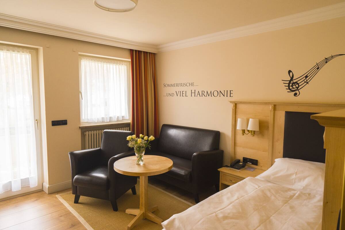 Una habitación de hotel con una cama, una mesita de noche de madera, un sillón de cuero negro, una pequeña mesa redonda con un jarrón de flores y una ventana con cortinas.