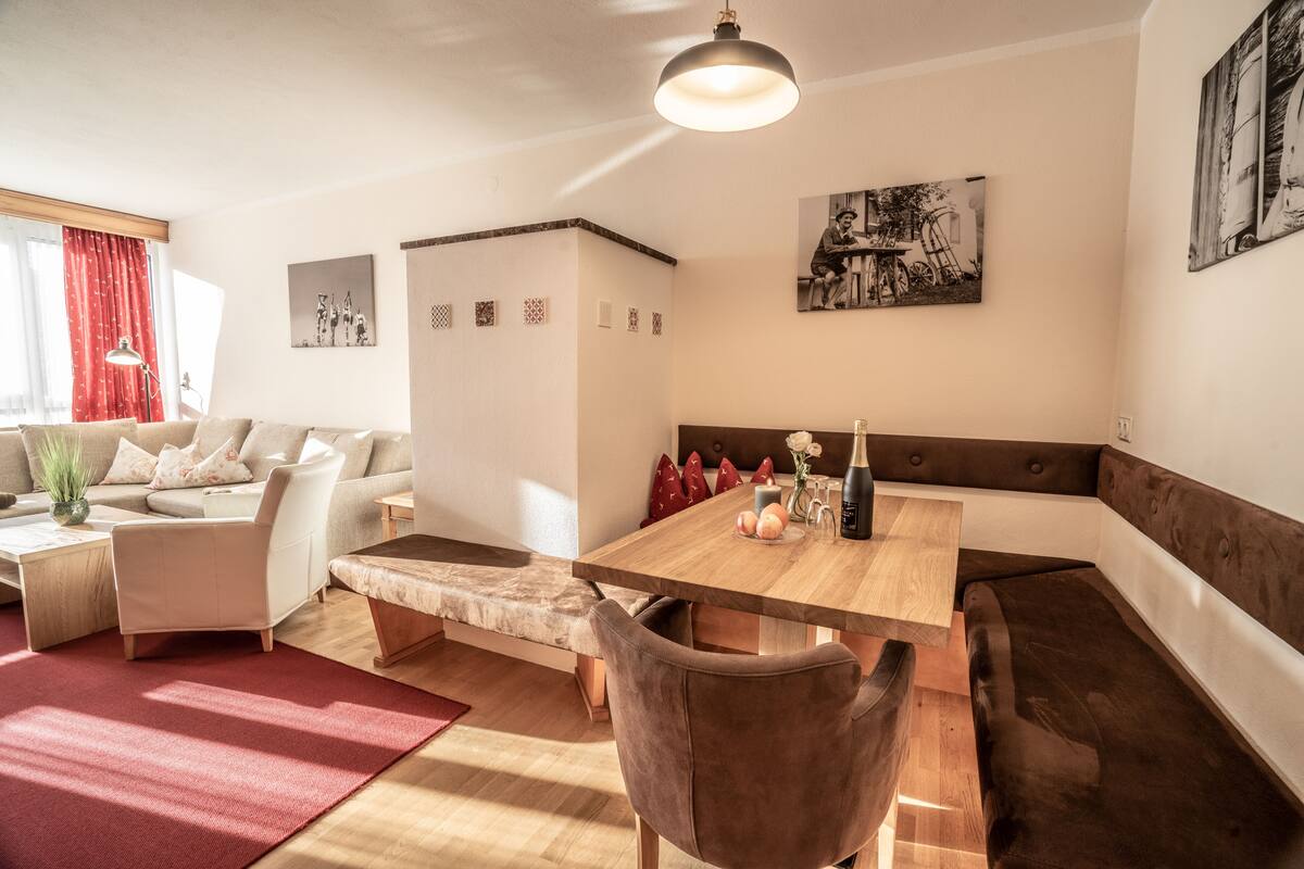 Una sala de estar moderna con una mesa de comedor de madera, un sofá y un banco. Hay cojines decorativos y un jarrón con flores sobre el sofá.
