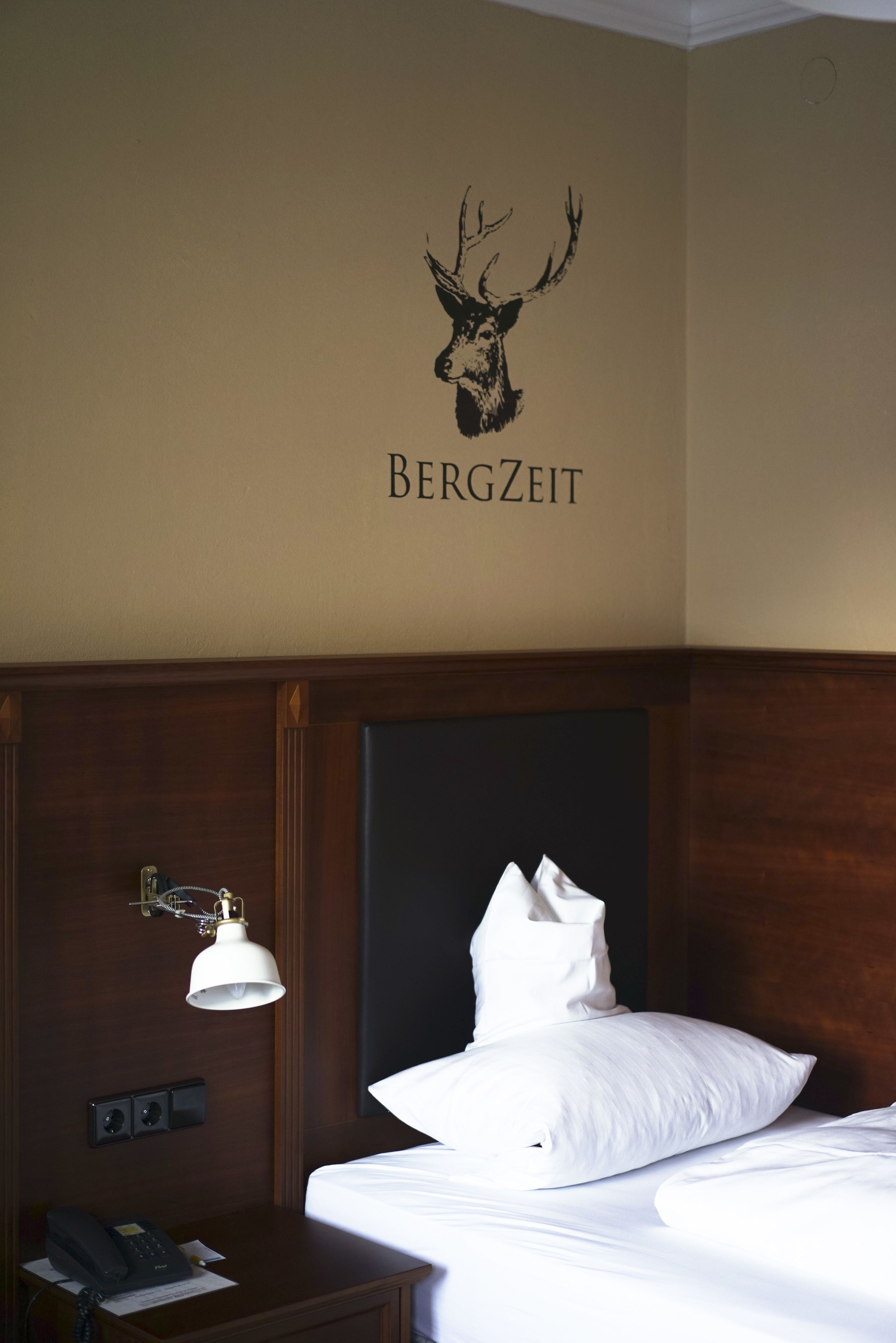 A hotel room with a bed, a nightstand, a lamp, and a wall decal of a deer head with the word "BERGZEIT".