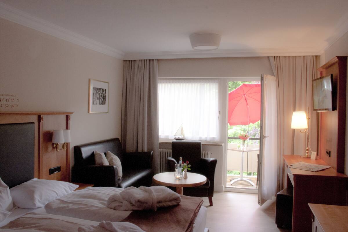 Habitación de hotel con cama, sofá, una mesita con un florero, una silla, televisor y balcón con sombrilla roja.
