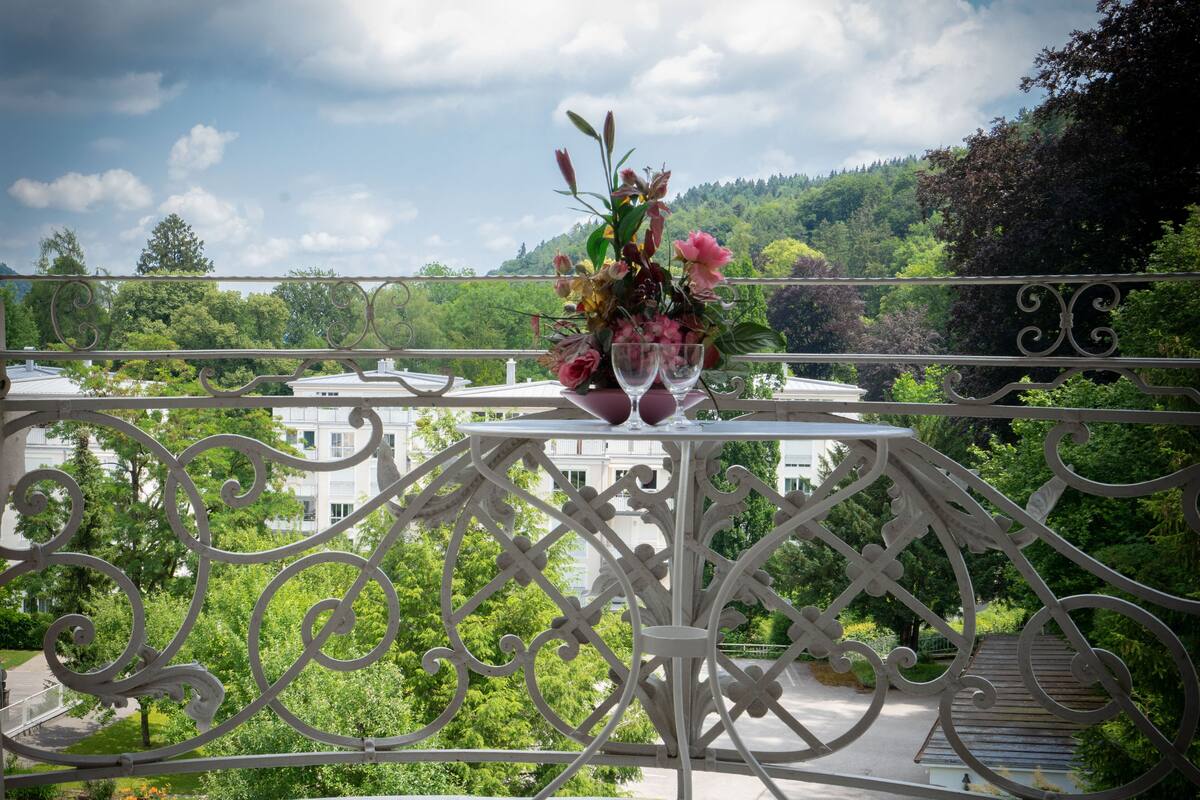 Un balcón con barandilla blanca, una mesa con un jarrón de flores y vista a un paisaje verde con árboles y edificios.