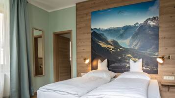 A hotel room with a bed, two bedside tables, a mirror, a poster of a mountain landscape, and a window with curtains.