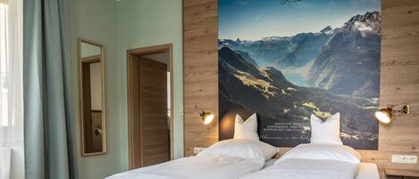 A hotel room with a bed, two bedside tables, a mirror, a poster of a mountain landscape, and a window with curtains.