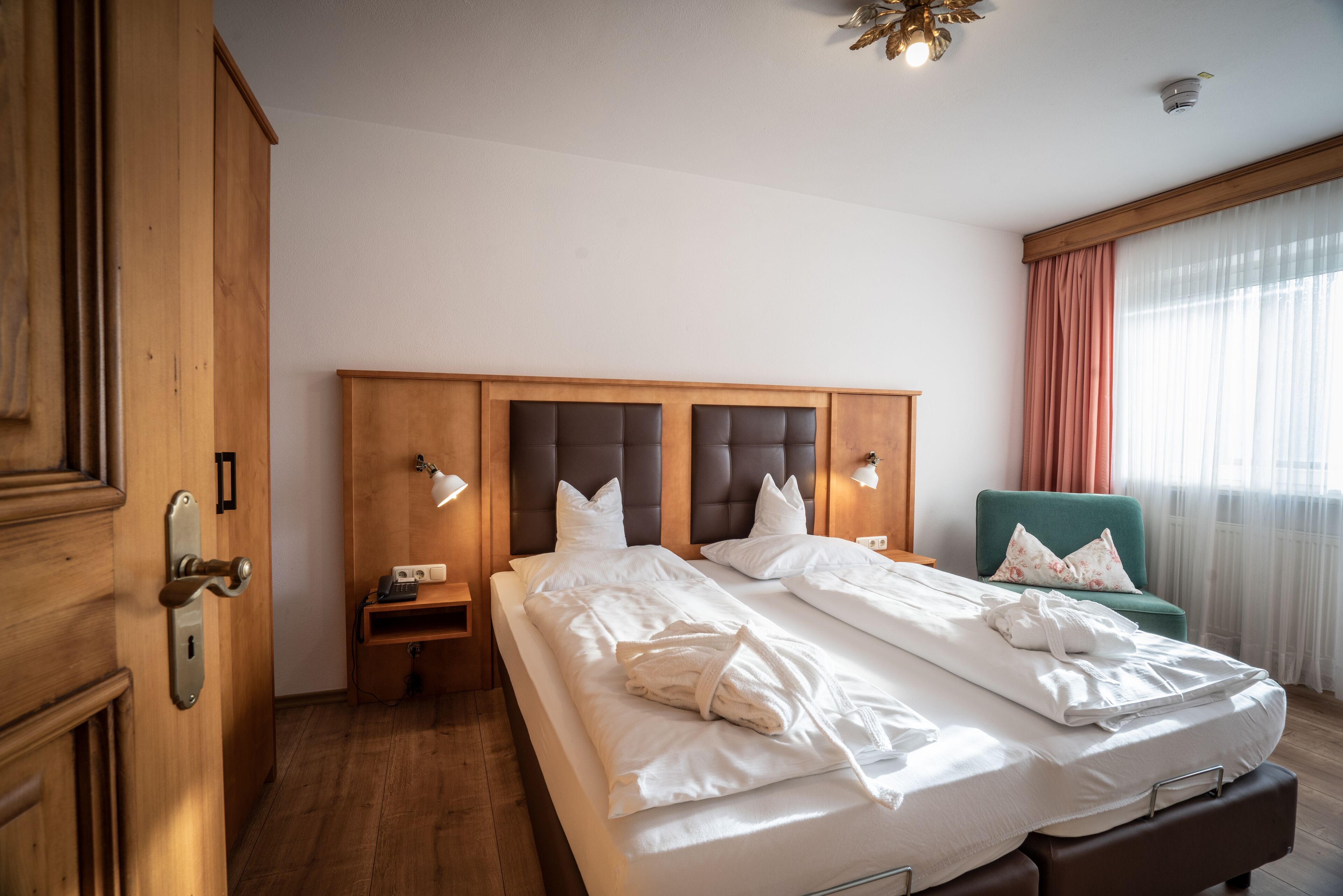 A hotel room with a large bed, wooden headboard, bedside lamps, a green armchair, and a wooden wardrobe.