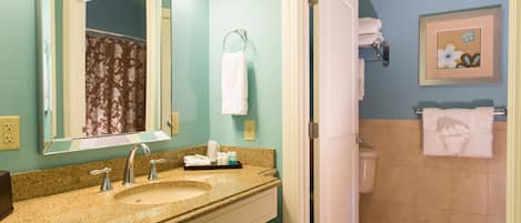 A bathroom with a sink, mirror, and toilet. There are towels hanging on the wall and a framed picture above the toilet.