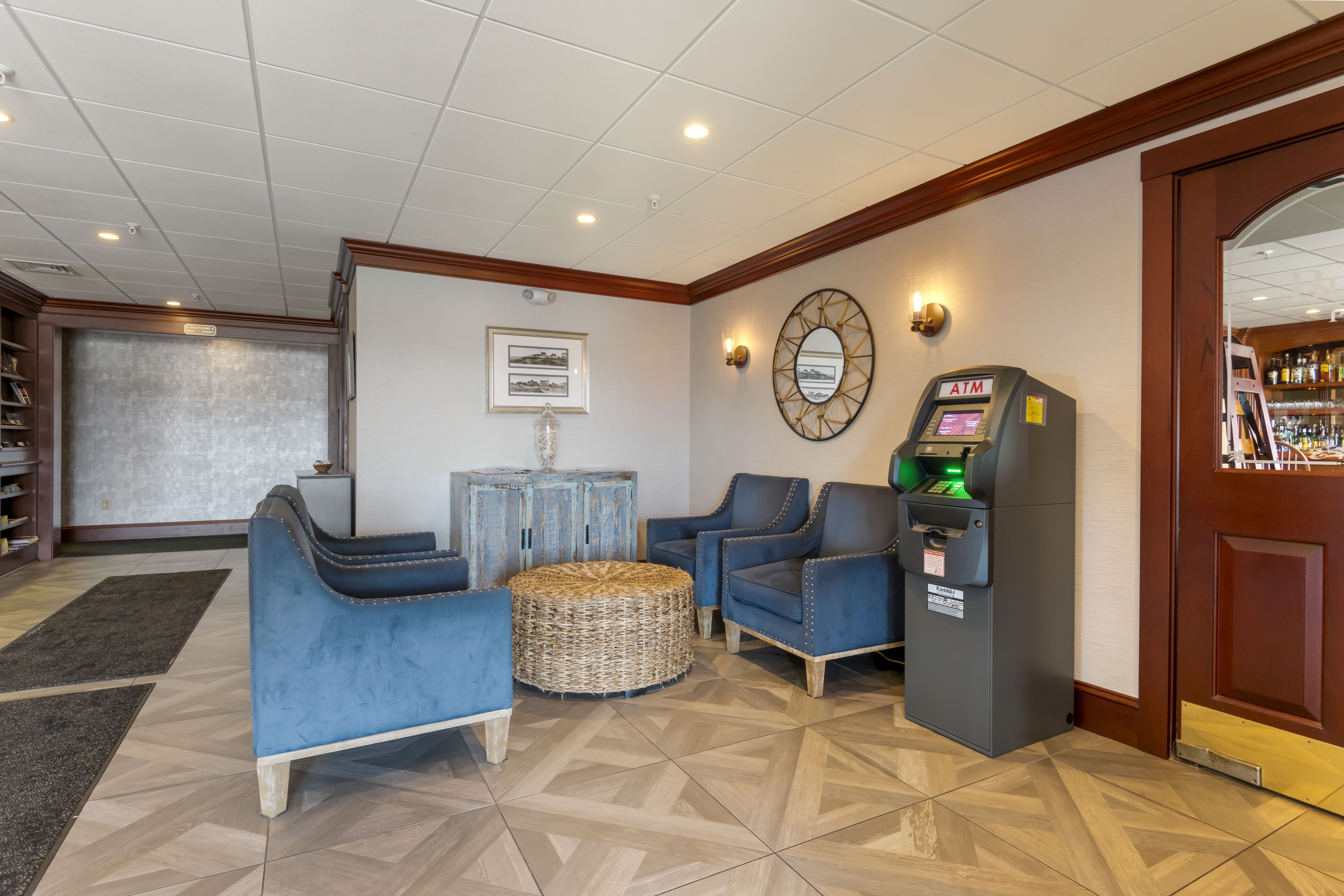 A lobby with a reception desk, blue armchairs, a wicker ottoman, an ATM, and a bookshelf.