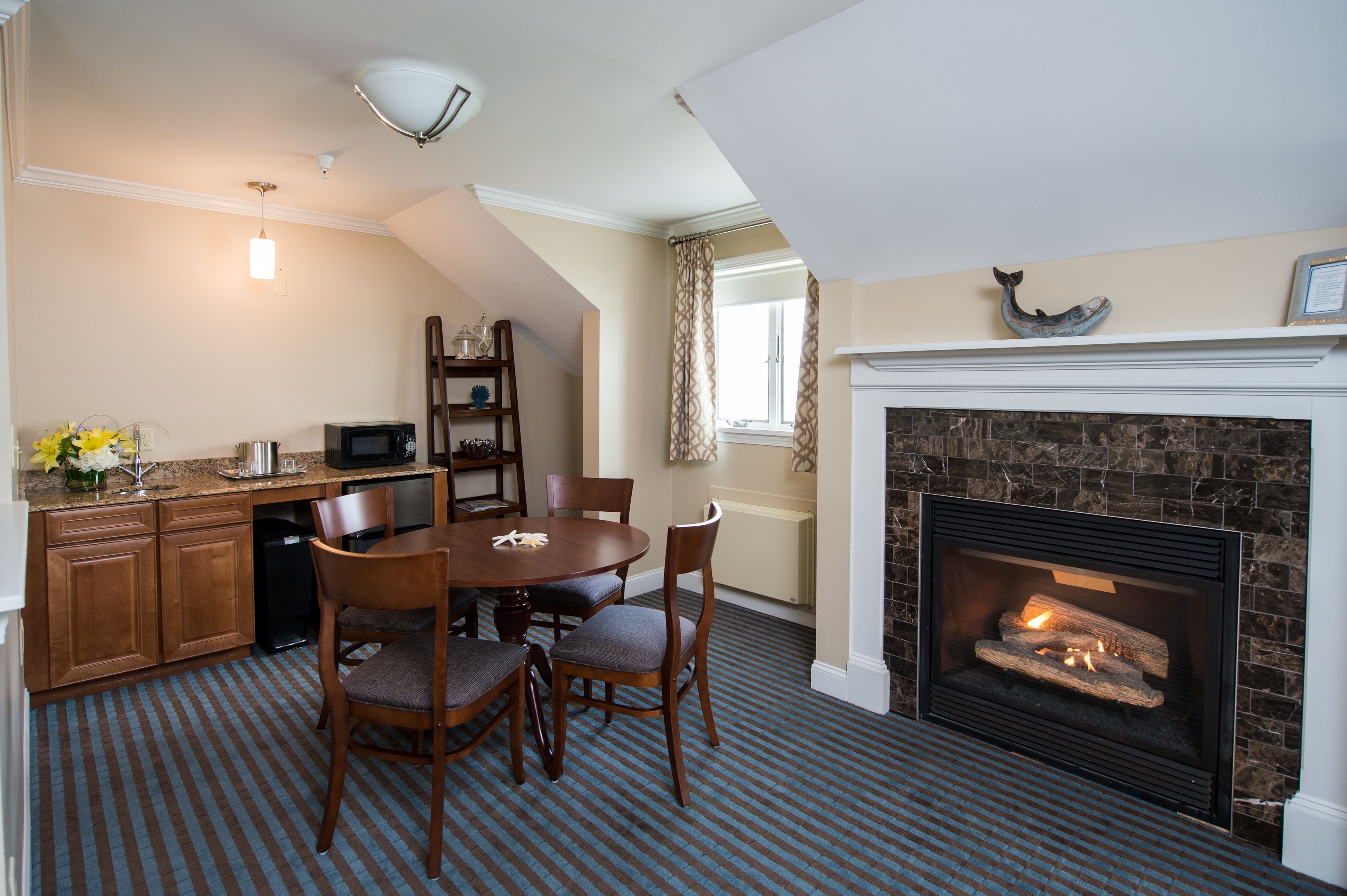 A dining area with a round table, chairs, a kitchen with cabinets, a microwave, and a fireplace with a fire.