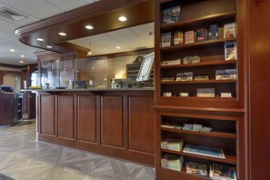 A reception desk with a wooden finish, a glass partition, and a digital information board. There are wooden shelves with magazines and brochures.